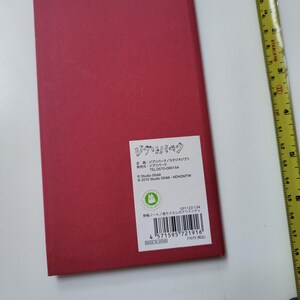 May include: A rectangular, red notebook with a white label featuring Japanese text and a Studio Ghibli logo. The notebook is on a white surface, with a yellow measuring tape in the background.