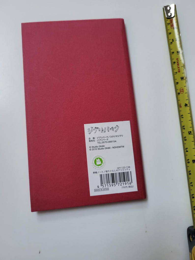 May include: A rectangular, red notebook with a white label featuring Japanese text and a Studio Ghibli logo. The notebook is on a white surface, with a yellow measuring tape in the background.