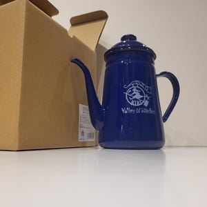 May include: A cobalt blue enamel teapot with a long spout and a handle. The teapot has a lid and a white graphic with the text "KIKI Valley of Witches". A cardboard box is partially visible in the background.