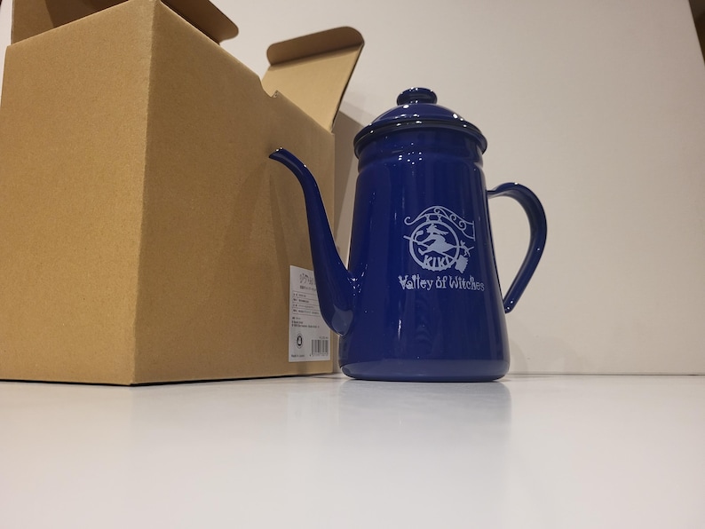 May include: A cobalt blue enamel teapot with a long spout and a handle. The teapot has a lid and a white graphic with the text "KIKI Valley of Witches". A cardboard box is partially visible in the background.