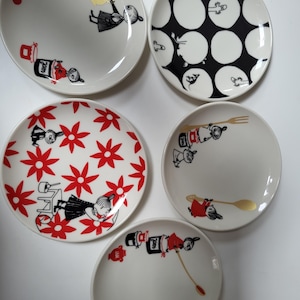 May include: A collection of five white ceramic plates with whimsical illustrations. Designs include red and black figures, red flowers, and black polka dots. Some plates feature gold accents, such as a fork or spoon.