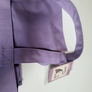 May include: A lavender tote bag with sturdy handles and a rectangular body. The bag has a tag attached with a cartoon illustration. The bag is made of a durable fabric, suitable for carrying various items.