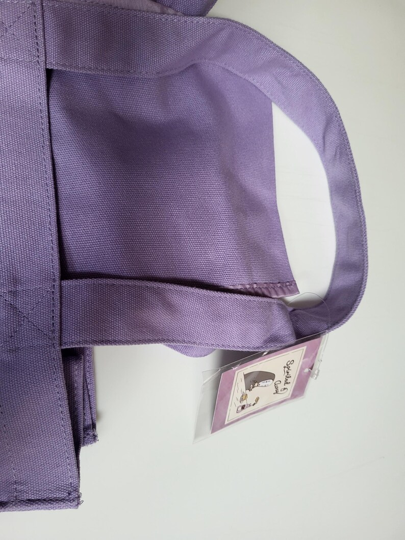 May include: A lavender tote bag with sturdy handles and a rectangular body. The bag has a tag attached with a cartoon illustration. The bag is made of a durable fabric, suitable for carrying various items.