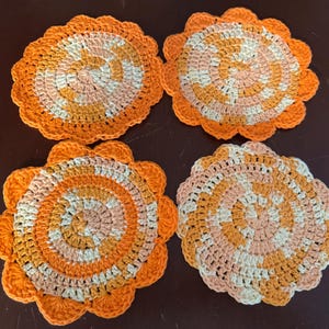 May include: Four crocheted coasters in a peach, white, and orange color scheme. The coasters have a circular design with a scalloped edge.