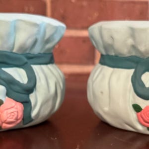 Peut inclure: Deux pots en céramique en forme de sacs avec un ruban bleu et des roses roses. Les pots sont de couleur bleu clair.