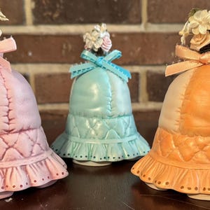 May include: Five porcelain bell figurines with floral accents and quilted fabric designs. The bells are in shades of blue, pink, orange, and lavender.
