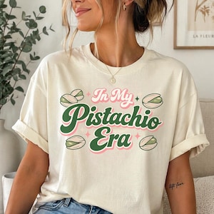 以下が含まれることがあります： クリーム色のTシャツ。テキスト「In My Pistachio Era」は緑とピンクで、ピスタチオのイラストと星のアクセントが特徴です。 シャツは袖をまくり、ゆったりとしたフィット感です。