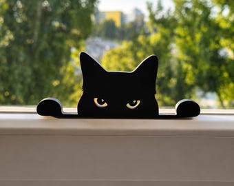 May include: A black cat silhouette cutout featuring pointed ears and two glowing yellow eyes. The cat figure is positioned horizontally with its paws extended to the sides, creating a flat, decorative piece with a sharp, clean edge profile.