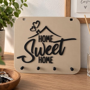 Könnte beinhalten: Ein rechteckiger, beigefarbener Wand-Schlüsselhalter mit dem schwarzen Schriftzug "HOME SWEET HOME" in einer Kombination aus Schreib- und Blockschrift. Das Design zeigt ein schwarzes Hausdach-Symbol und zwei kleine Herzen über dem Text.