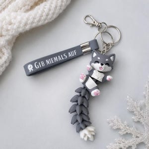 Könnte beinhalten: Ein grauer Schlüsselanhänger mit einem Silikonband, das den weißen Schriftzug GIB NIEMALS AUF und ein kleines Emblem trägt. Daran befestigt ist eine bewegliche, grau-weiße fuchsähnliche Figur mit rosa Pfoten, einem lächelnden Gesicht und einem langen, segmentierten Schwanz.