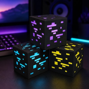 Könnte beinhalten: Drei schwarze würfelförmige Lampen mit pixelartigen Ausschnitten. Der obere Würfel strahlt violettes Licht aus, der untere linke Würfel leuchtet hellblau und der untere rechte Würfel strahlt gelbes Licht durch die rechteckigen Öffnungen.