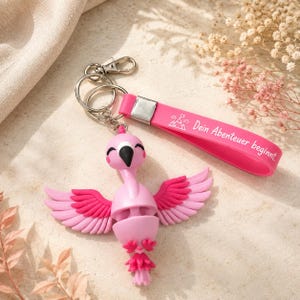 Könnte beinhalten: Ein rosa Flamingo-Schlüsselanhänger mit einem flexiblen Silikonband, das den weißen Text "Dein Abenteuer beginnt." trägt. Der Flamingo-Anhänger hat ausgebreitete Flügel mit geschichteten rosa Federn, einen schwarzen Schnabel und einen Schwanz.