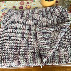 May include: A hand-knitted blanket in shades of purple, gray, and pink. The chunky knit blanket is folded, showcasing the texture and color variations. The blanket is made of a soft, plush yarn.