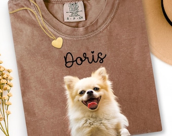 Op de afbeelding: Een bruin t-shirt met de naam "Doris" in zwart script boven een lachende, lichtgekleurde hond. Een gouden hartvormige hangerketting en een beige hoed zijn ook zichtbaar.