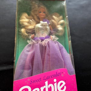 Puede incluir: Muñeca Barbie Sweet Lavender en su embalaje original. La muñeca tiene pelo largo rubio y viste un vestido lavanda con un corpiño blanco. La caja es rosa y presenta el logotipo de Barbie.