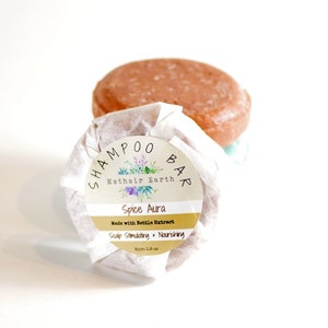 May include: A brown shampoo bar wrapped in white paper with a label that reads "SHAMPOO BAR Mathair Earth Spice Aura Made with Nettle Extract Scalp Stimulating + Nourishing Aprx. 2.8 oz"
