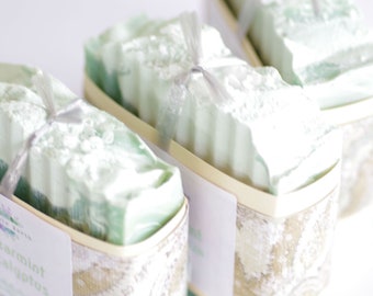 May include: Three bars of handmade soap wrapped in white and silver ribbon. The soap is a light green color and has a white swirl design. The soap is labeled "Spearmint Eucalyptus".