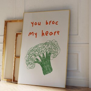 Könnte beinhalten: Ein weißer gerahmter Druck mit dem Text "you broc my heart" in Orange und Grün. Unter dem Text befindet sich eine grüne Strichzeichnung eines Brokkolis.