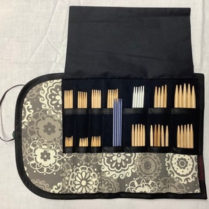 May include: A black and gray floral patterned knitting needle case, open to display various sizes of wooden and silver knitting needles. The case is rolled up and secured with a black strap.