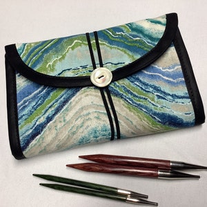 May include: A fabric knitting needle case with a blue, green, and white marbled pattern, black trim, and a button closure. Two pairs of knitting needles are visible: one pair is dark red, and the other is green.
