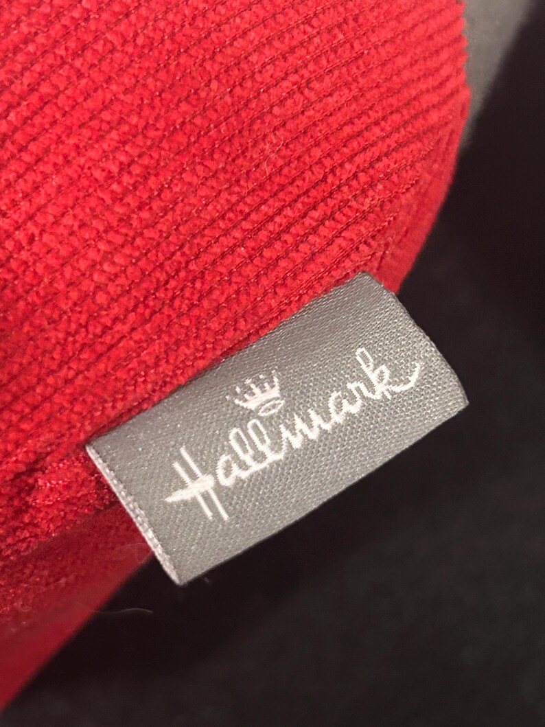 May include: A red fabric pillow with a gray tag that says "Hallmark" with a crown above the word.