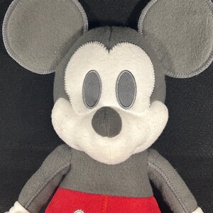 May include: A gray and white plush Mickey Mouse toy with red shorts and white buttons. The toy has a stitched smile and black eyes.