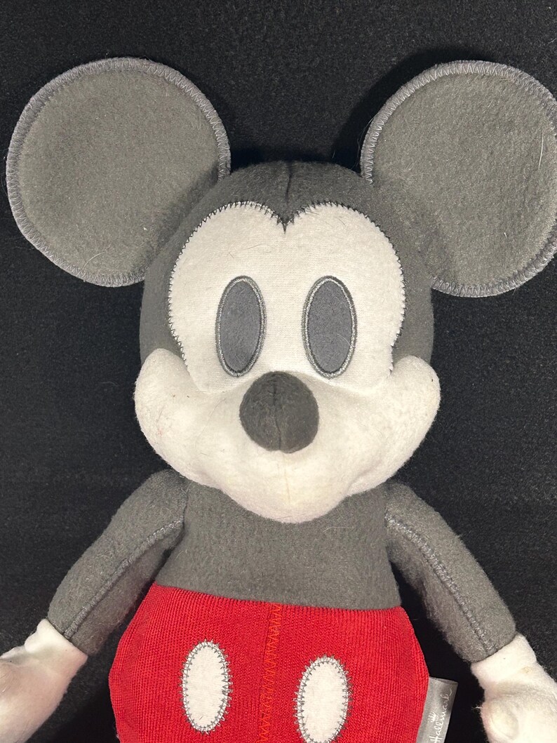 May include: A gray and white plush Mickey Mouse toy with red shorts and white buttons. The toy has a stitched smile and black eyes.