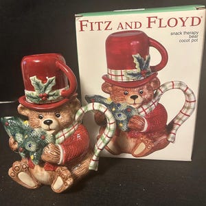 May include: Two ceramic bear-shaped teapots with red top hats and matching jackets. Each bear holds a Christmas tree and wears a plaid scarf. The box behind the teapots reads "Fitz and Floyd".