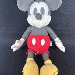 May include: A gray plush Mickey Mouse toy with red shorts and yellow shoes. The toy has a white face with black eyes and a black nose.