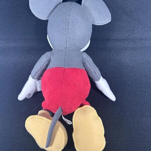 May include: A plush Mickey Mouse toy with gray fur, red shorts, and yellow shoes. The toy is sitting on a black surface.