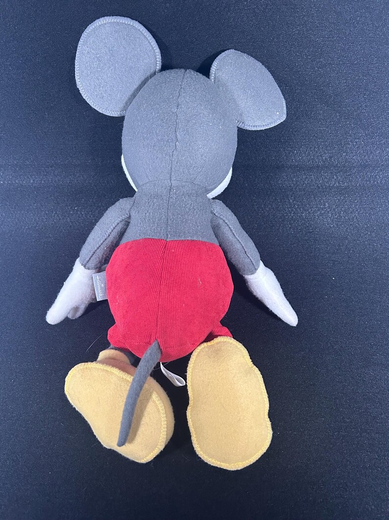 May include: A plush Mickey Mouse toy with gray fur, red shorts, and yellow shoes. The toy is sitting on a black surface.