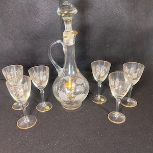 May include: A set of six clear glass wine glasses with gold rims and a matching clear glass decanter with a gold rim and stopper. The glasses and decanter are etched with a floral design.