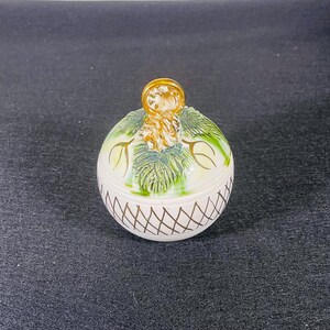 May include: A white ceramic Christmas ornament shaped like a ball with a gold-colored pine cone design and a gold-colored loop on top.