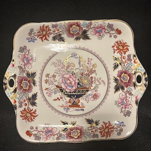 May include: A rectangular, off-white ceramic serving tray with a floral design. The tray features a central floral arrangement in a dark blue vase, surrounded by a circular border and floral patterns. Gold trim and handles complete the design.
