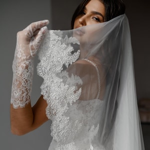 May include: A white wedding veil with detailed lace edging. The veil is draped over a white wedding dress, and the image also shows white lace gloves. The text "PrincessVeil" is visible on the dress.