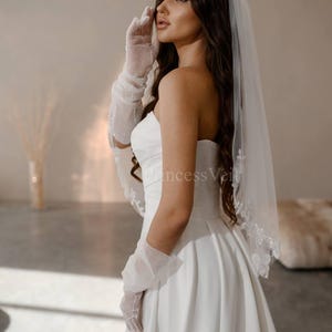 May include: A bride wearing a white strapless wedding dress with a white veil and gloves. The veil has a beaded edge.