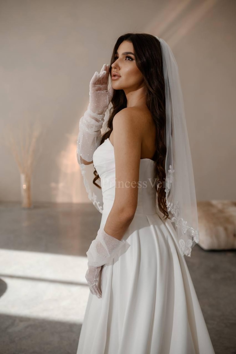 May include: A bride wearing a white strapless wedding dress with a white veil and gloves. The veil has a beaded edge.