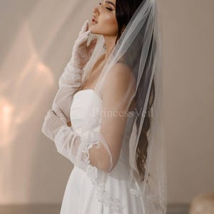 May include: A bride wearing a white wedding dress with a long, white veil. The veil has a delicate lace trim and is adorned with small, sparkling crystals. The bride is wearing white gloves and has her hair pulled back.