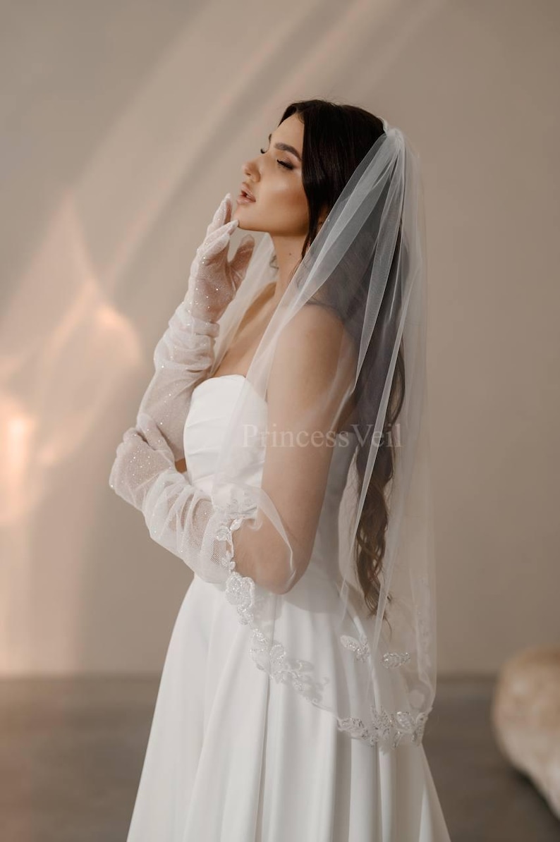 May include: A bride wearing a white wedding dress with a long, white veil. The veil has a delicate lace trim and is adorned with small, sparkling crystals. The bride is wearing white gloves and has her hair pulled back.