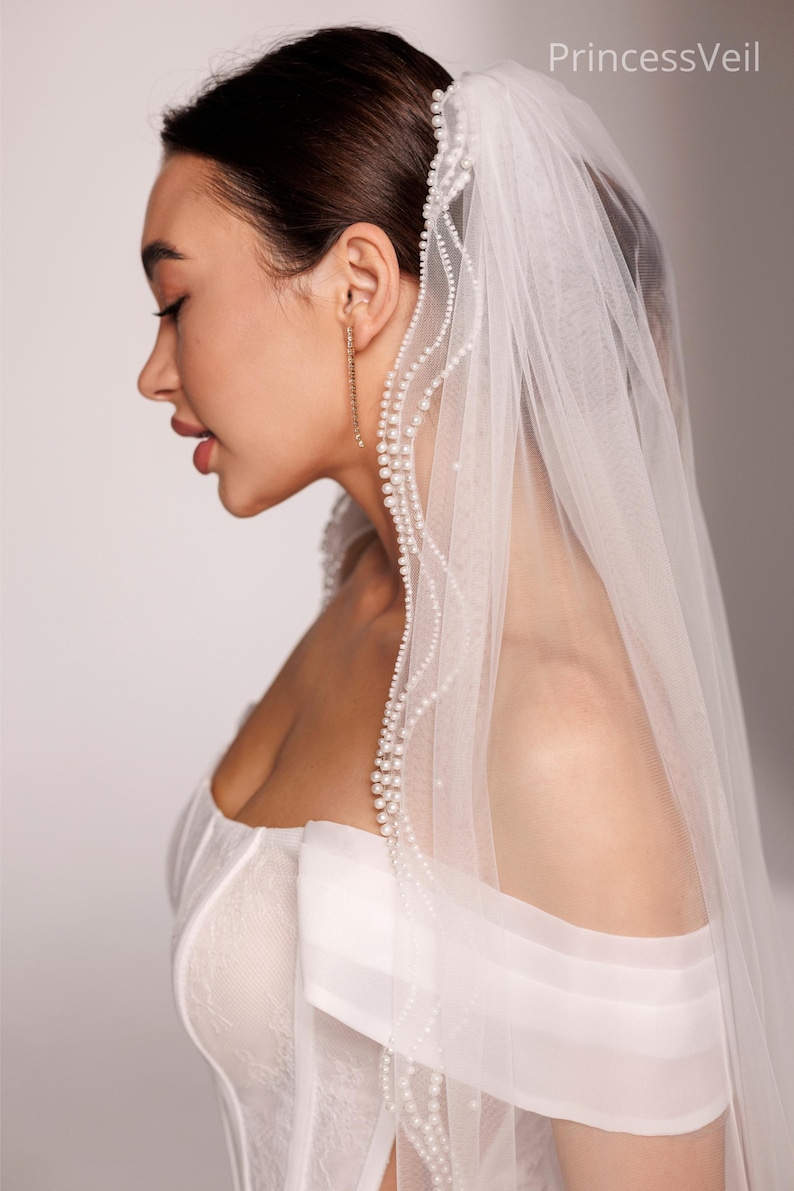 May include: A side profile of a person wearing a white wedding dress and veil. The veil is adorned with a delicate pearl trim. The text "PrincessVeil" is visible in the upper right corner.