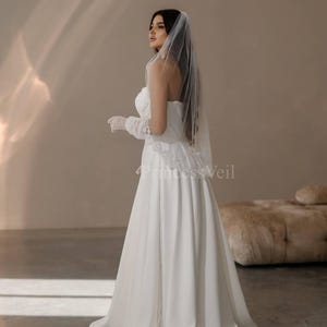 May include: A bride wearing a white strapless wedding dress with a long veil. The dress has a fitted bodice and a flowing skirt. The bride is standing in a room with a light brown background.