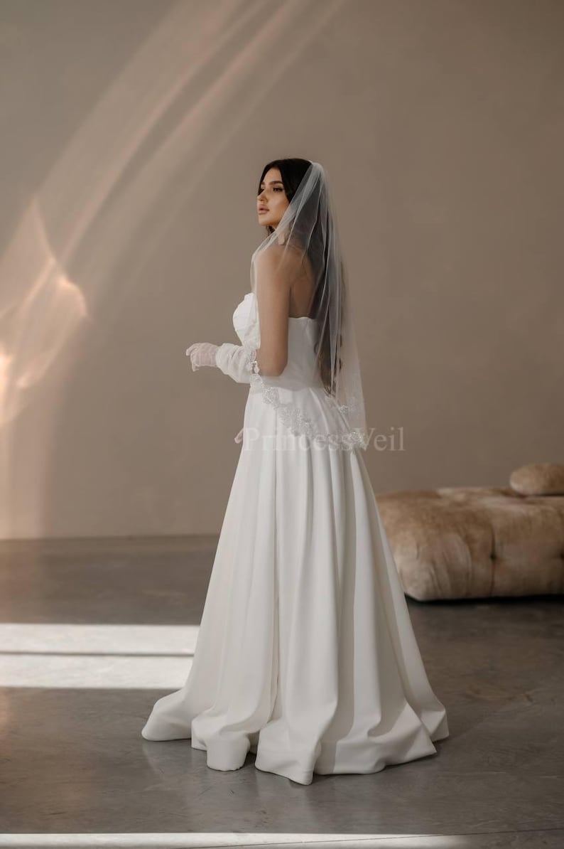 May include: A bride wearing a white strapless wedding dress with a long veil. The dress has a fitted bodice and a flowing skirt. The bride is standing in a room with a light brown background.