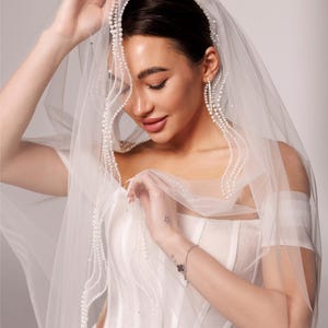 May include: A sheer white bridal veil with delicate pearl accents. The veil is draped over a white wedding dress. The text "PrincessVeil" is visible in the upper right corner. The veil is held up by a person.
