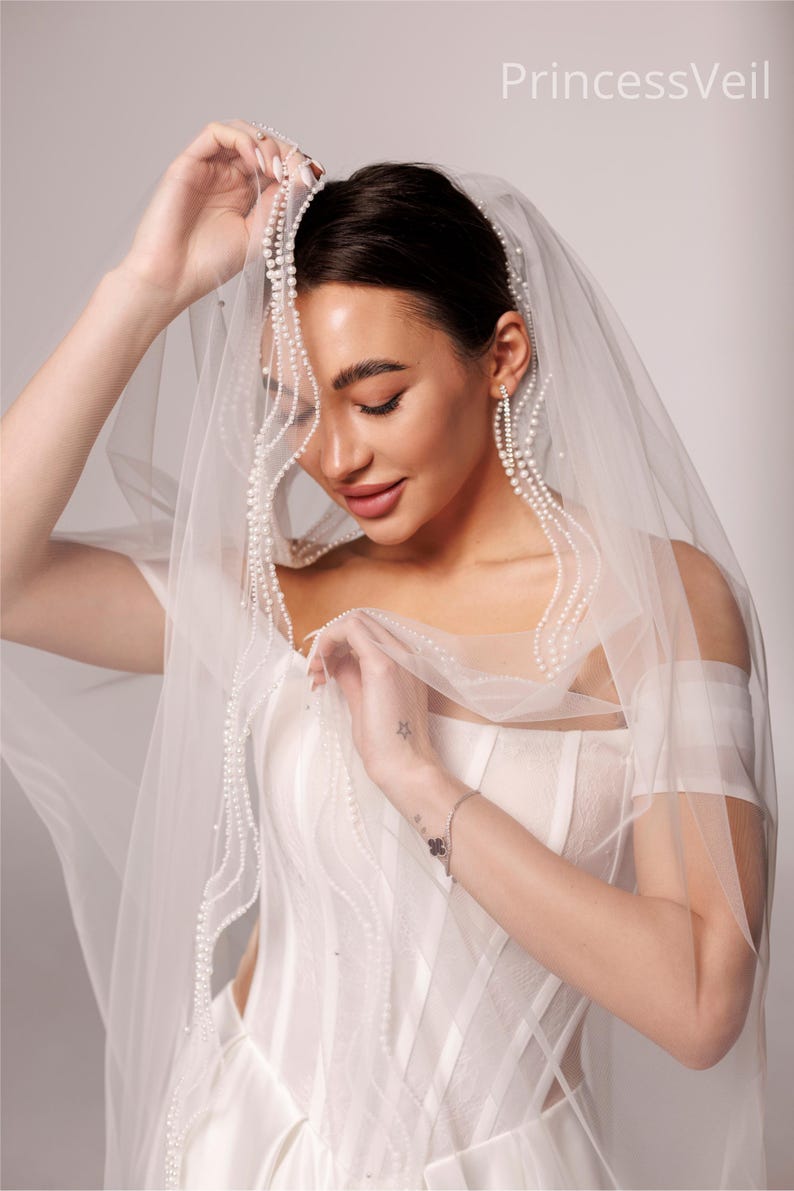May include: A sheer white bridal veil with delicate pearl accents. The veil is draped over a white wedding dress. The text "PrincessVeil" is visible in the upper right corner. The veil is held up by a person.