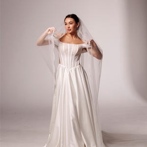 May include: A white satin wedding dress with a corset bodice and a flowing skirt. The bride is holding a sheer veil. The text "PrincessVeil" is in the upper right corner.