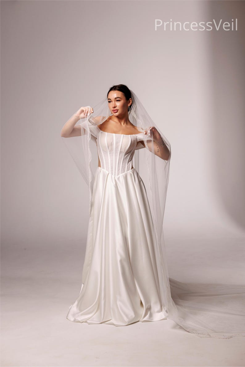 May include: A white satin wedding dress with a corset bodice and a flowing skirt. The bride is holding a sheer veil. The text "PrincessVeil" is in the upper right corner.