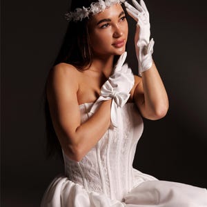 May include: A white strapless corset top with lace detailing, paired with white satin gloves featuring small bows at the wrists and a white floral headpiece. The text PrincessVeil is visible in the upper right corner of the image.
