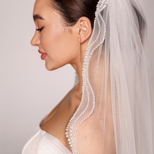 May include: A sheer white bridal veil with a delicate pearl trim. The veil is draped over the shoulder and head, with the text "PrincessVeil" visible in the upper right corner. The model is wearing gold earrings.