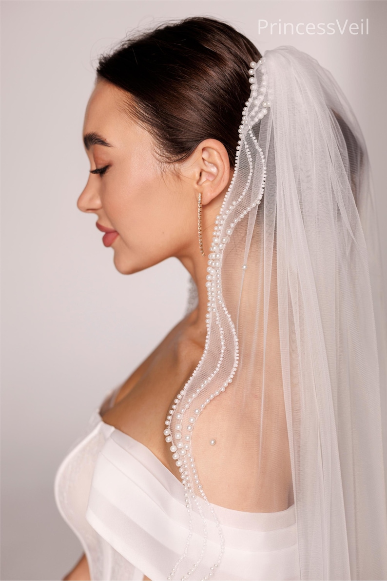 May include: A sheer white bridal veil with a delicate pearl trim. The veil is draped over the shoulder and head, with the text "PrincessVeil" visible in the upper right corner. The model is wearing gold earrings.
