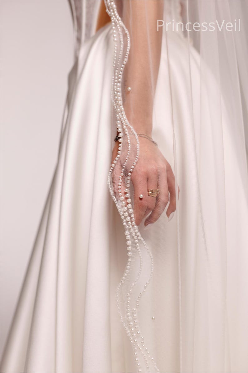 May include: A white bridal veil with a delicate pearl trim. The veil is held by a hand wearing a ring. The text "PrincessVeil" is visible on the veil.